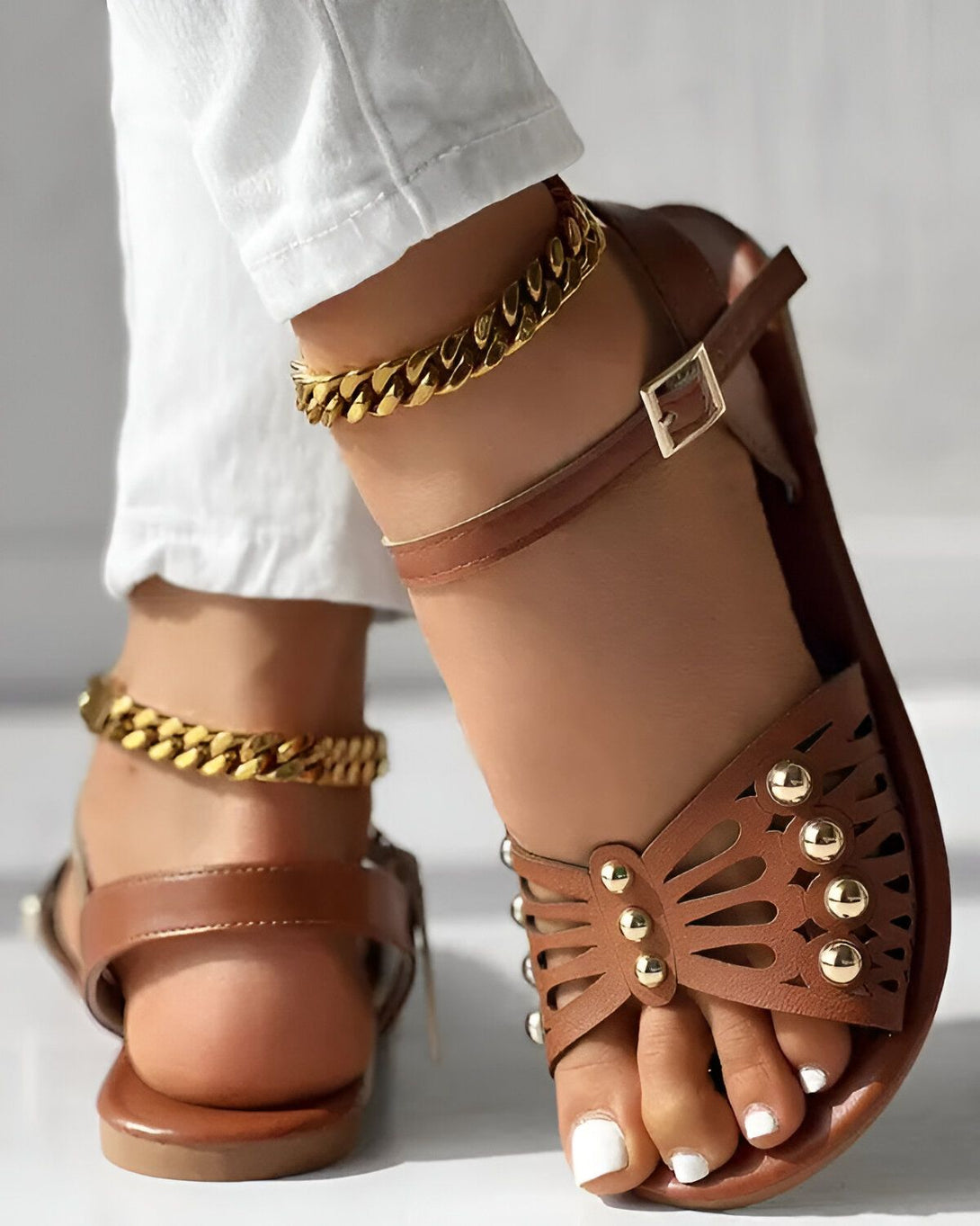 Une femme porte des sandales marron avec des détails dorés. Elle porte également un pantalon blanc et des bracelets de cheville en or.
