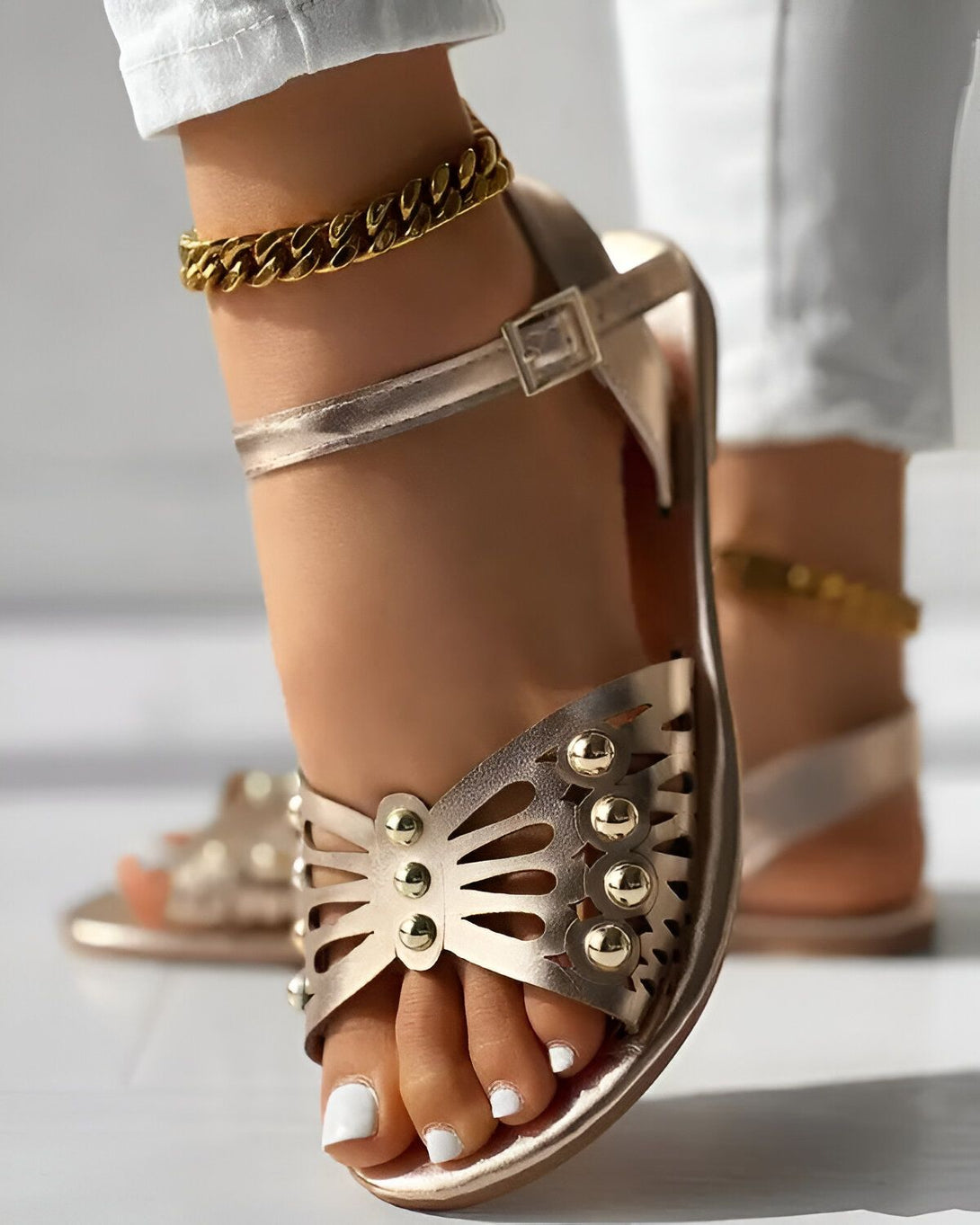 Une femme porte des sandales dorées à talons hauts. Les sandales sont en cuir et ont un design unique avec des découpes en forme de papillon sur le dessus. La femme a les ongles des pieds peints en blanc et porte un bracelet de cheville en or.