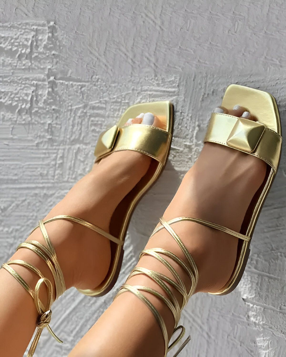 Une femme porte des sandales dorées à talons hauts. Les sandales sont attachées aux chevilles par des lacets fins. La femme est assise sur un mur et ses jambes sont croisées.