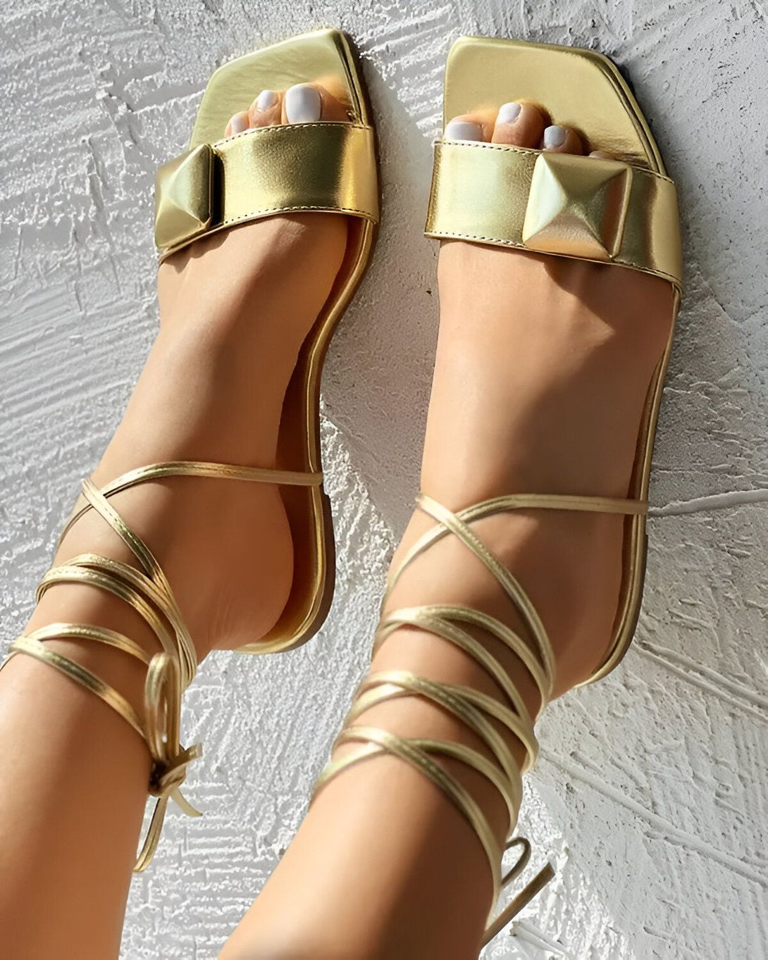 Une femme porte des sandales dorées à talons hauts. Les sandales sont en cuir et ont des brides qui s'enroulent autour de la cheville. La femme a les ongles des pieds peints en blanc.