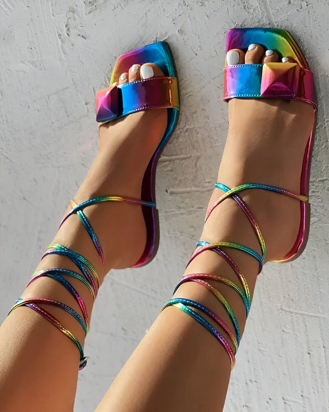 Une femme porte des sandales à talons hauts multicolores. Les sandales sont en cuir et ont des brides qui s'enroulent autour de la cheville. La femme a les ongles des pieds peints en blanc.