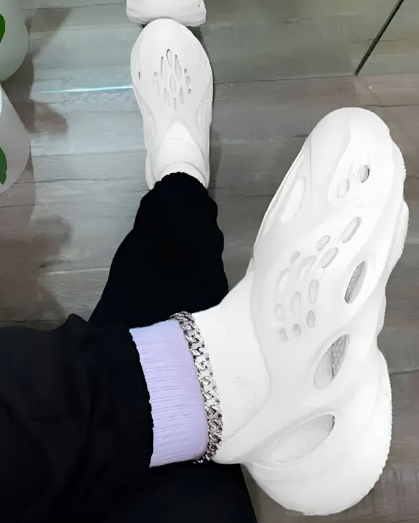 Une personne porte des chaussures blanches trouées avec un pantalon noir et des chaussettes mauves.
