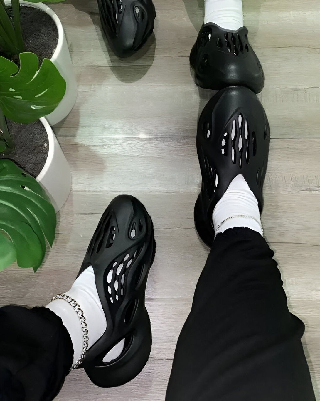 Une personne porte des sandales noires trouées avec des chaussettes blanches. Il est assis sur un sol en bois. Il y a des plantes vertes à côté de lui.