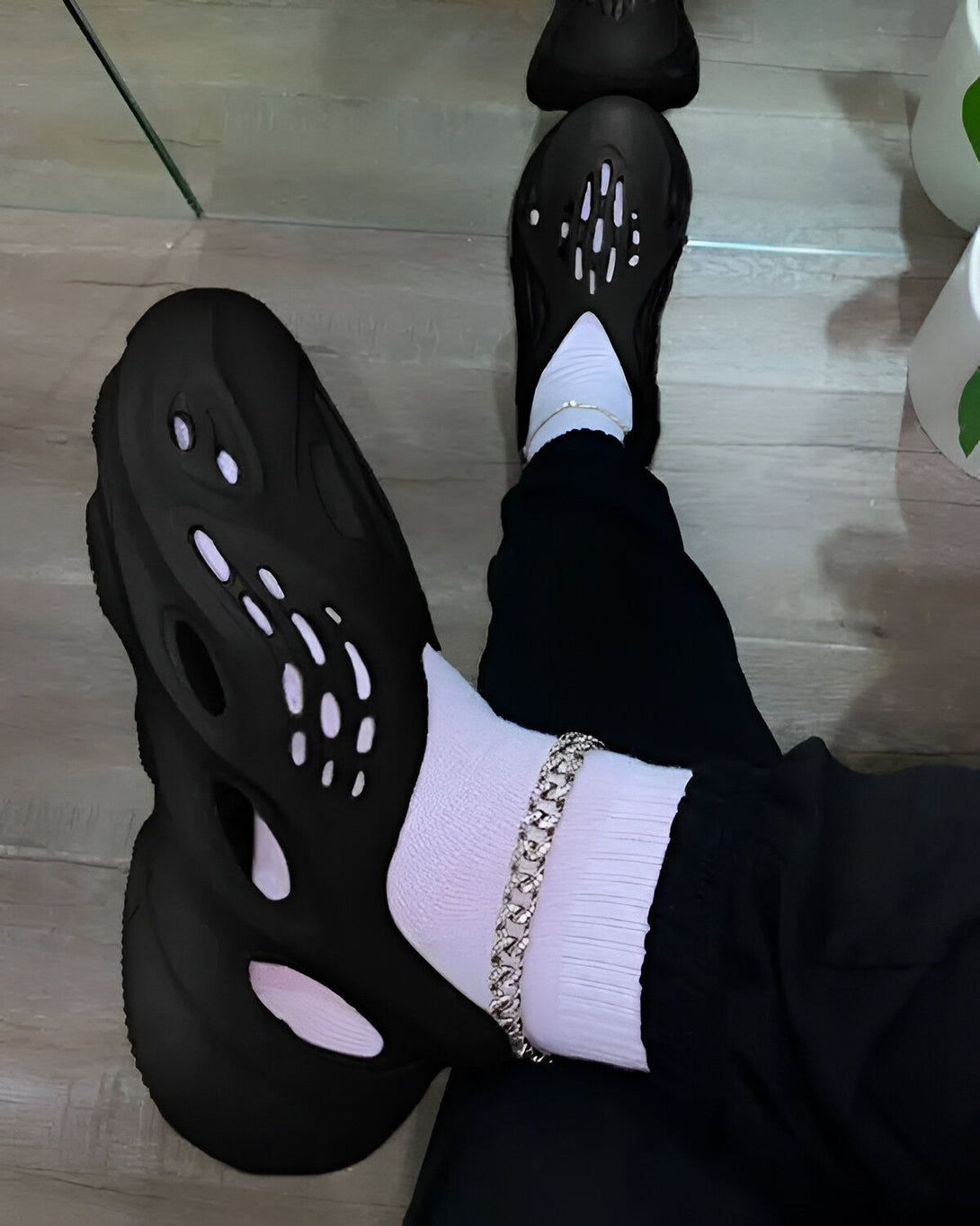 Une personne porte des chaussettes blanches, un pantalon noir et des chaussures noires trouées. Il y a aussi un reflet de la personne dans le miroir.