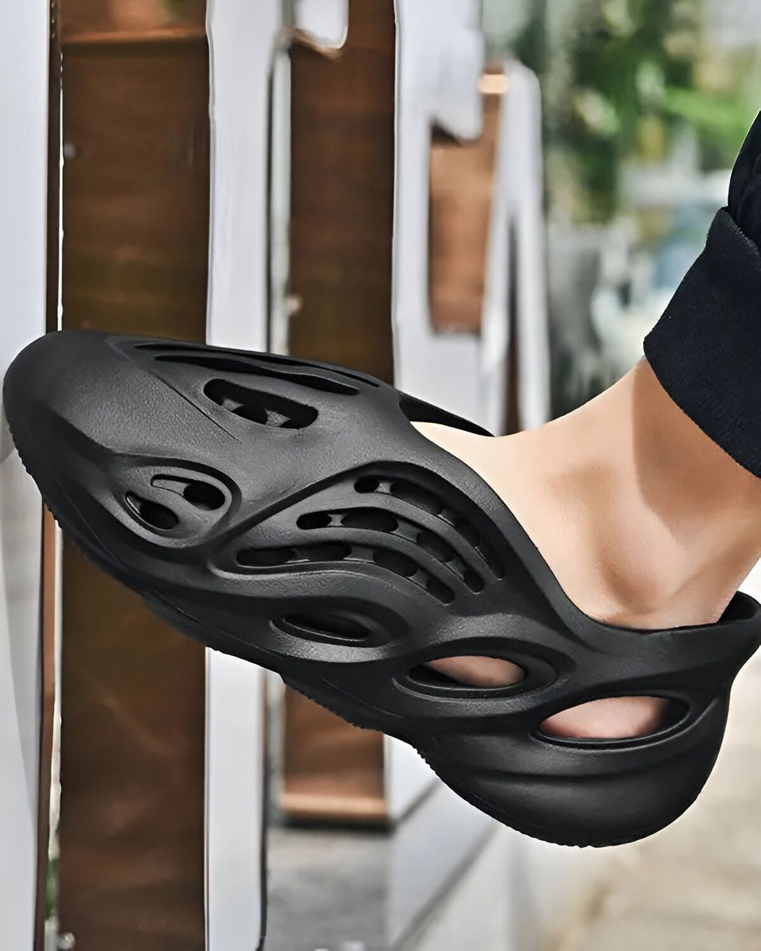 Une personne porte des sandales noires ajourées. Les sandales sont en plastique et ont une semelle épaisse. La personne porte des chaussettes noires.
