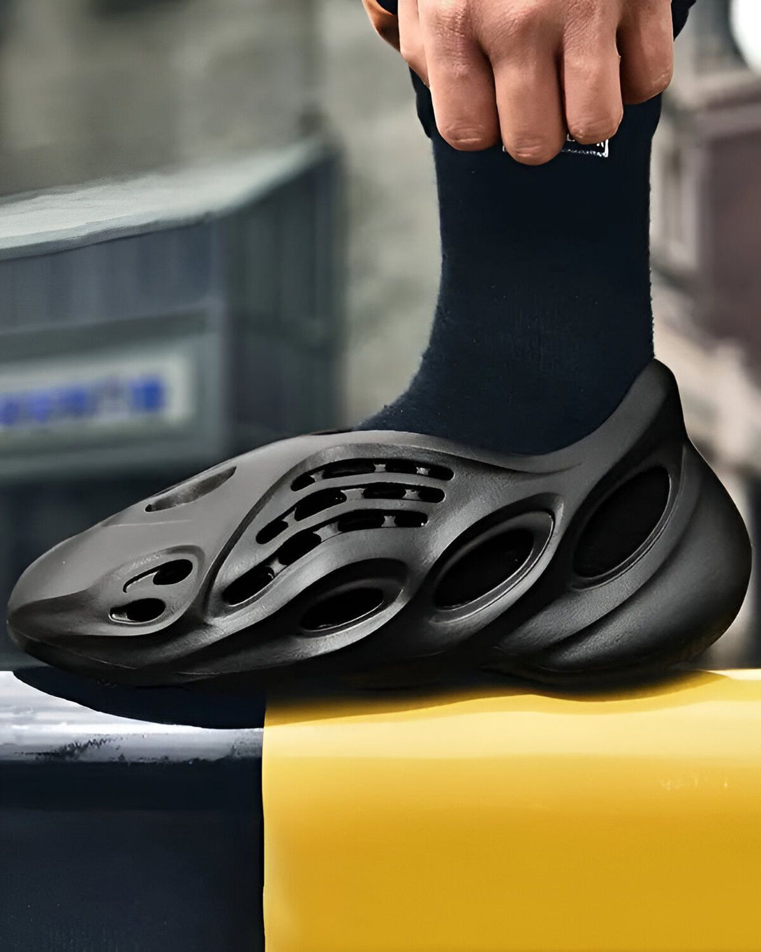 Une personne porte des chaussures noires avec des chaussettes noires. Les chaussures sont en mousse et ont des trous. La personne est debout sur une surface jaune.