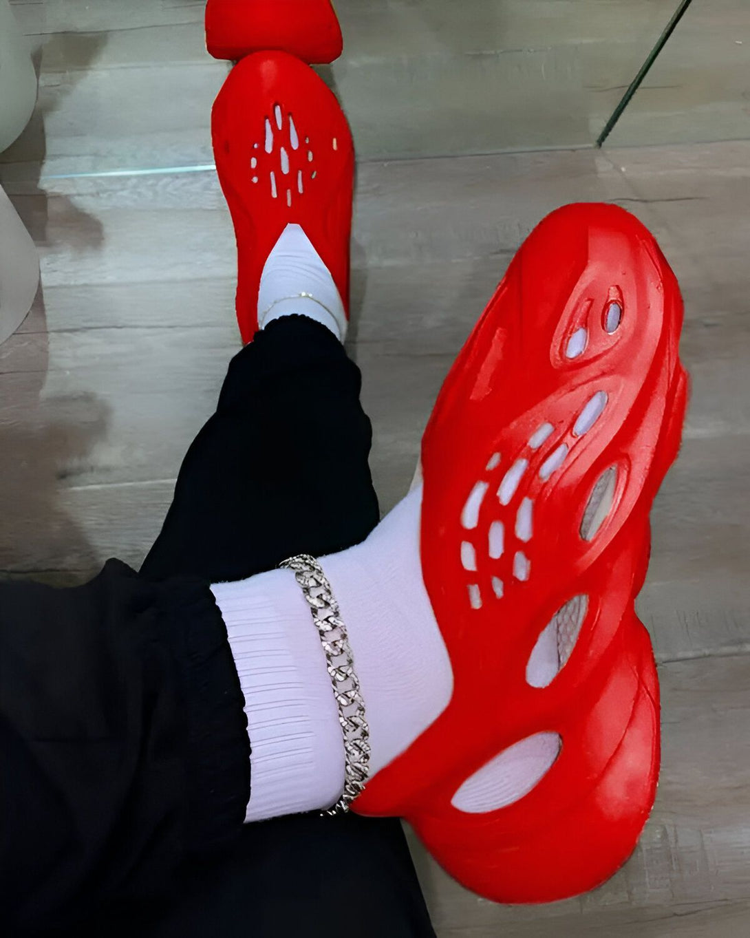 Une personne porte des chaussettes blanches et des sandales rouges trouées. La personne porte également un pantalon noir et une chaîne en argent à la cheville.