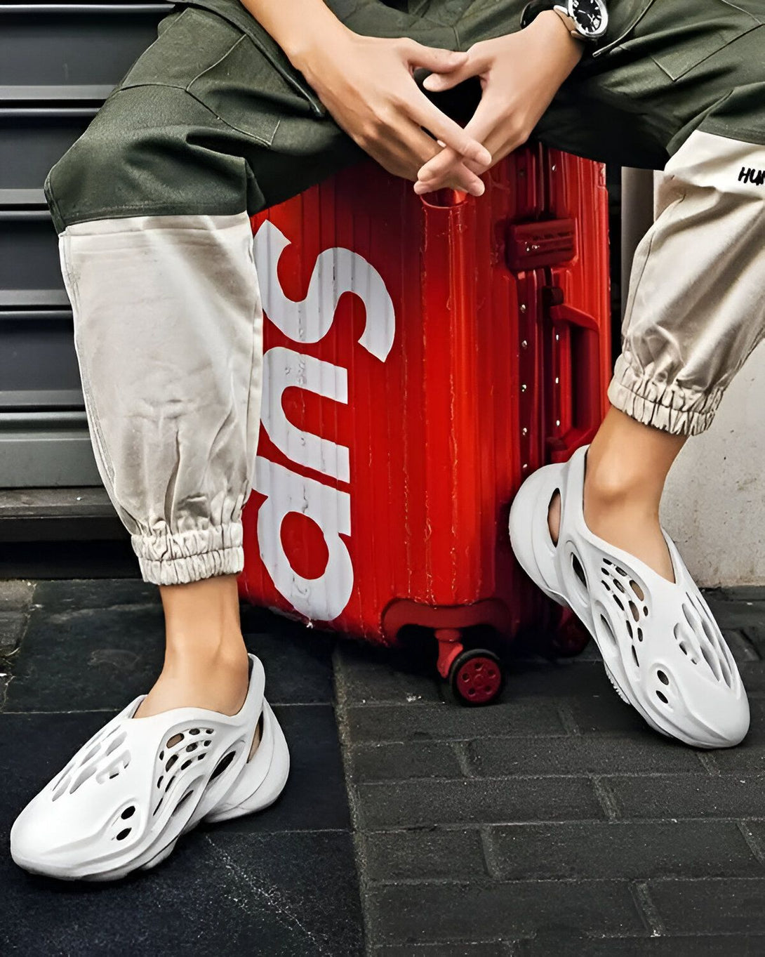 Une personne porte des sandales blanches trouées avec un pantalon cargo beige et vert et est assise sur une valise rouge.