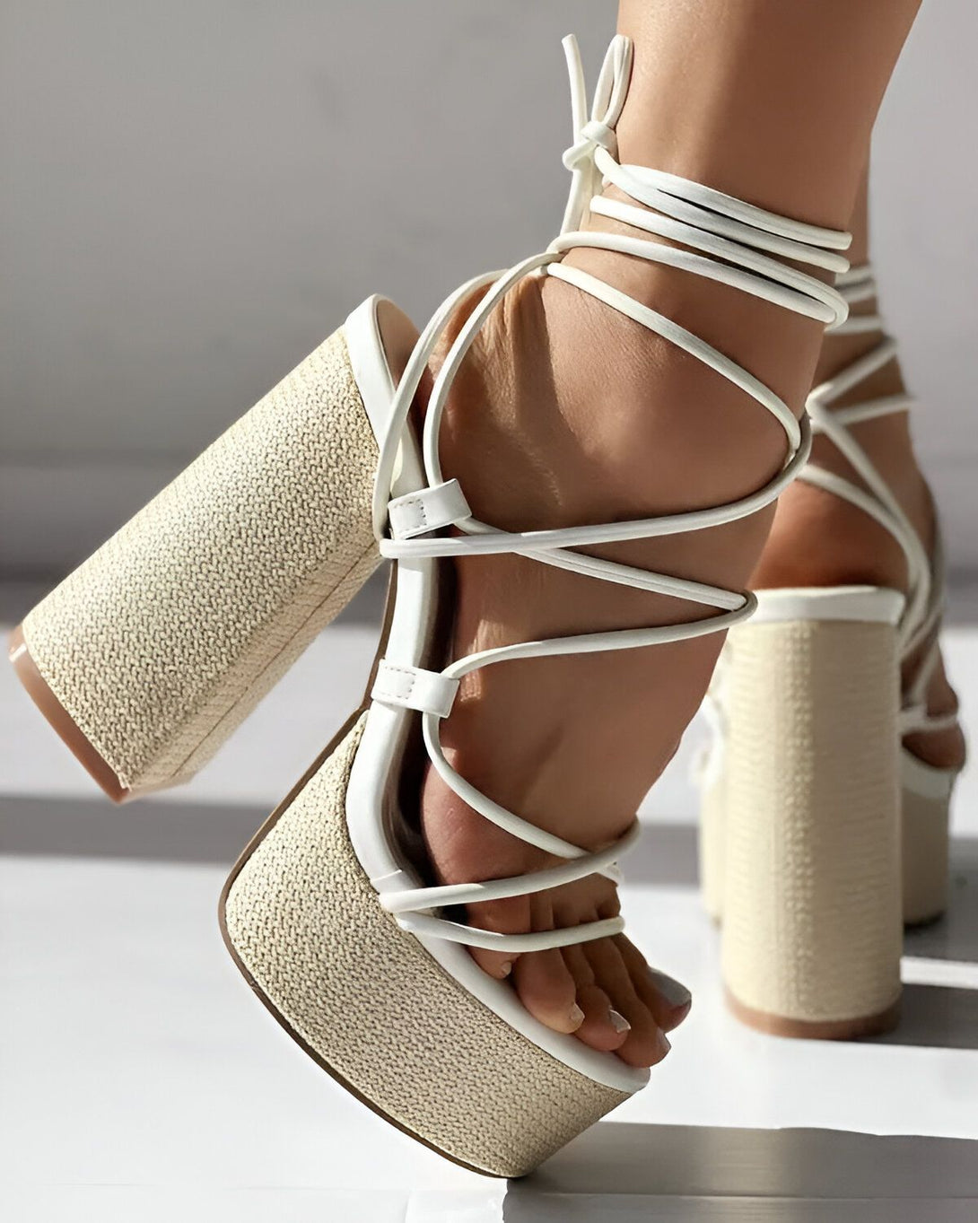 Une image d'une femme portant des sandales à talons hauts blancs. Les sandales sont à plateforme et ont des brides qui s'enroulent autour de la cheville. La femme est debout sur un sol blanc.