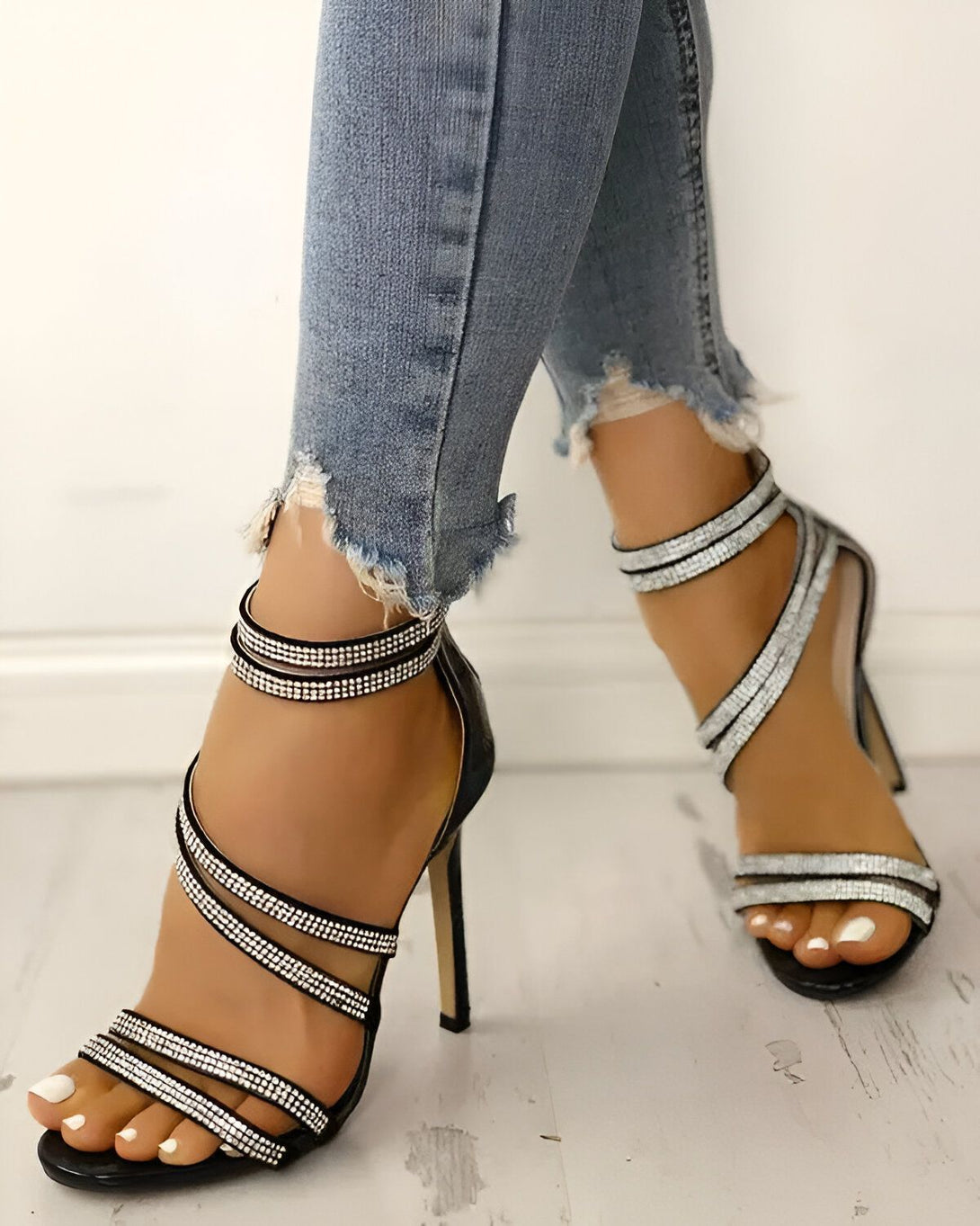 Une femme porte des sandales à talons hauts noires avec des brides à strass. Elle porte également un jean bleu déchiré.