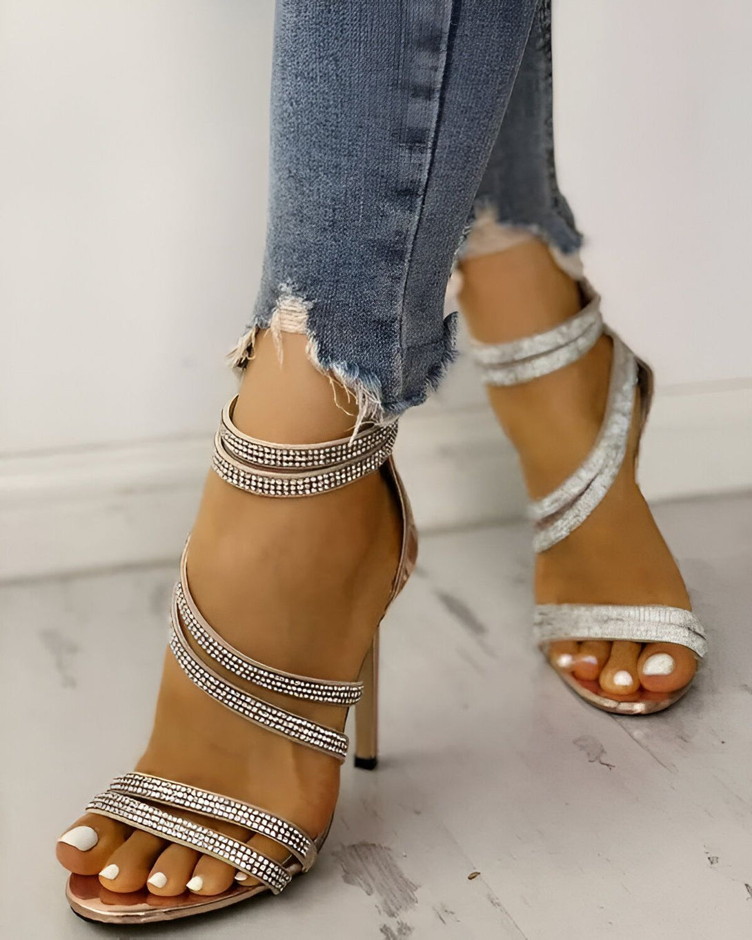 Une femme porte des sandales à talons hauts argentées. Les sandales sont ornées de strass. La femme porte un jean bleu déchiré.