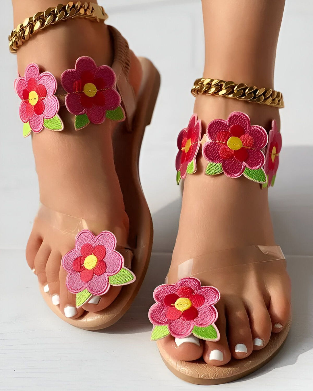 Une femme porte des sandales plates transparentes à fleurs roses. Les sandales sont ornées de fleurs en tissu rose et vert. La femme a les ongles des pieds peints en blanc. Elle porte également une chaîne dorée à la cheville.