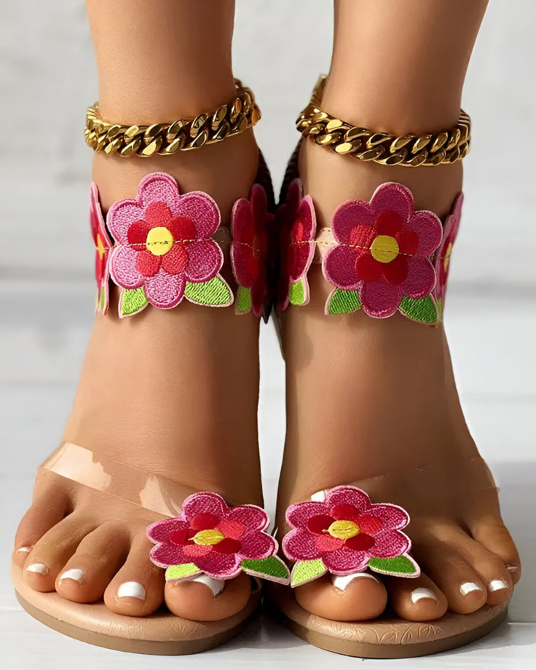 Une paire de sandales à talons hauts transparentes avec des fleurs roses brodées sur le dessus et une chaîne dorée autour de la cheville.