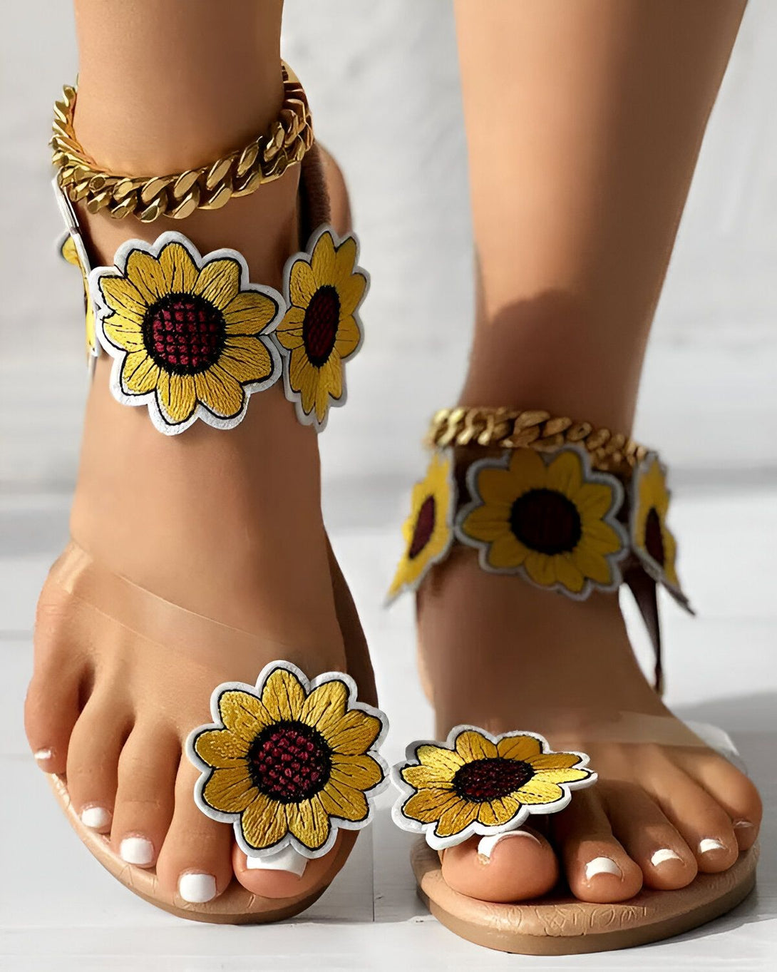 Une femme porte des sandales plates à enfiler avec des fleurs de tournesol brodées sur le dessus et une chaîne dorée autour de la cheville.