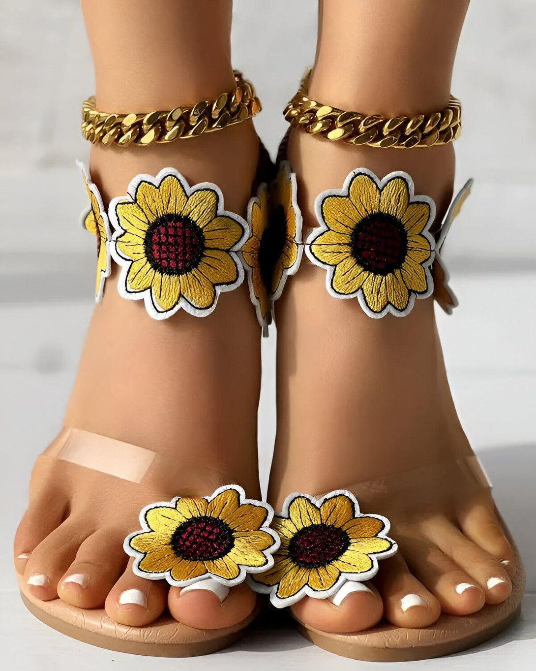Une paire de sandales plates transparentes à talons hauts avec des fleurs de tournesol jaunes et rouges brodées sur le dessus et une chaîne dorée autour de la cheville.
