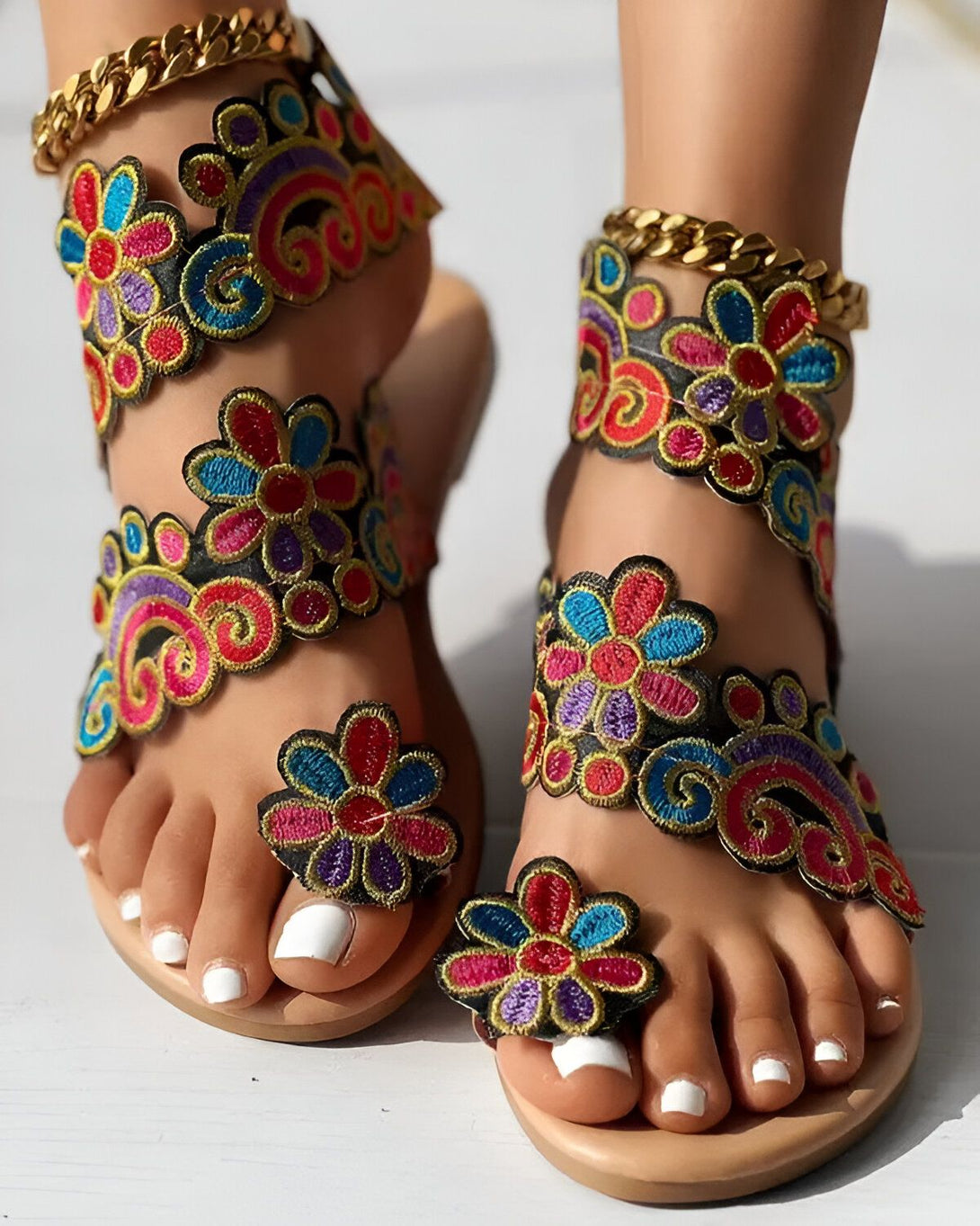 Une femme porte des sandales plates brodées de fleurs colorées. Les sandales sont attachées aux chevilles par des chaînes dorées.