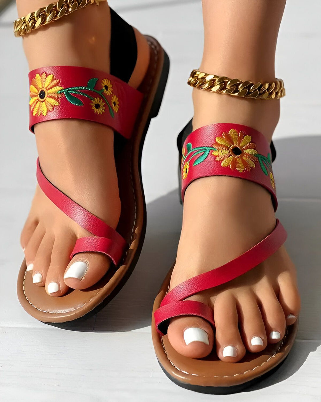 Une paire de sandales plates rouges avec des fleurs jaunes brodées sur le dessus. Les sandales sont portées par une femme qui a les ongles des pieds peints en blanc.