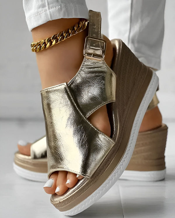 Une femme porte des sandales compensées dorées. Les sandales sont en cuir et ont une bride à boucle. La femme porte un jean blanc et un chemisier blanc. Elle a les ongles des pieds peints en blanc.