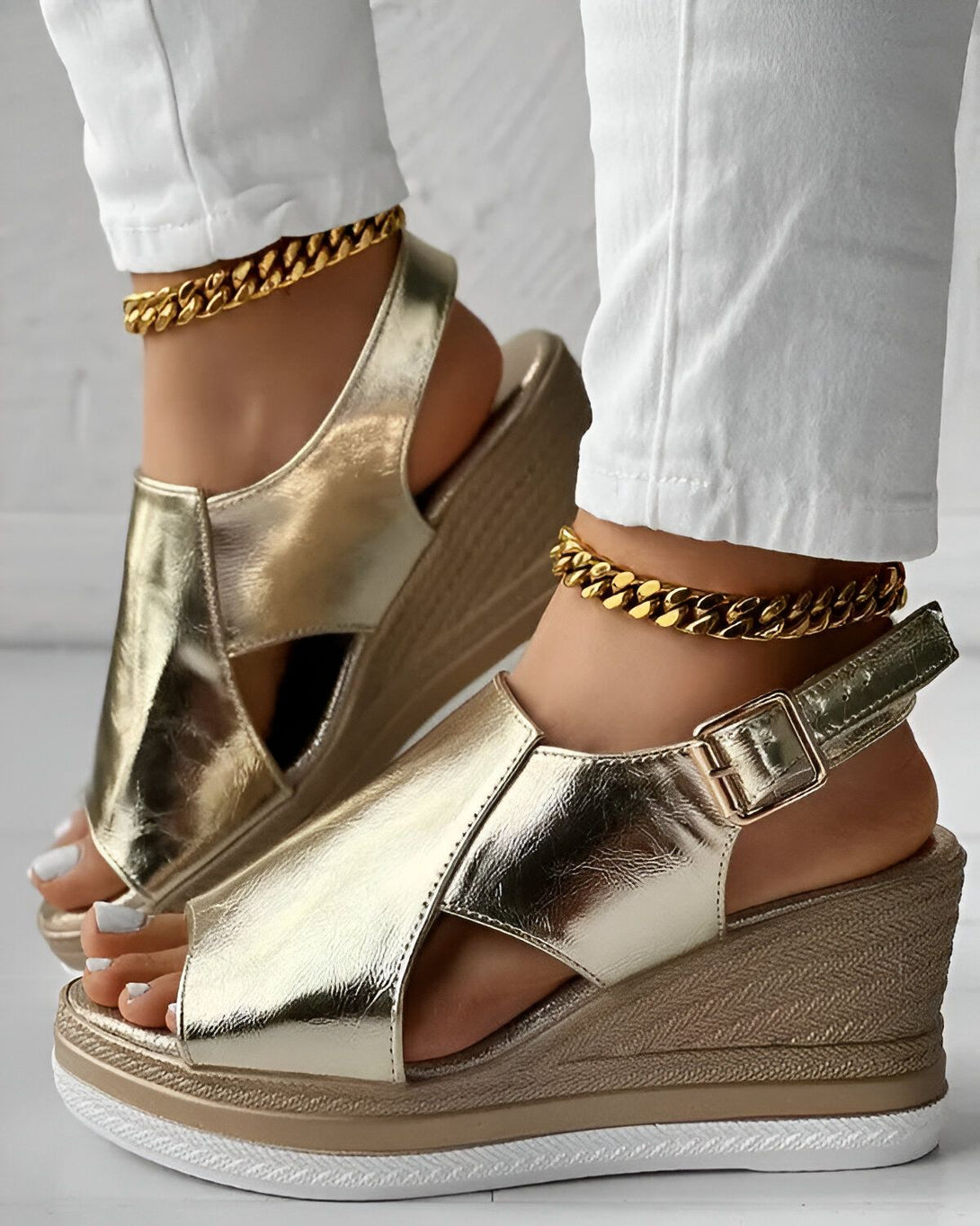 Une femme porte des sandales compensées dorées. Les sandales sont en cuir et ont une bride à boucle. La femme porte également un jean blanc et un chemisier blanc.