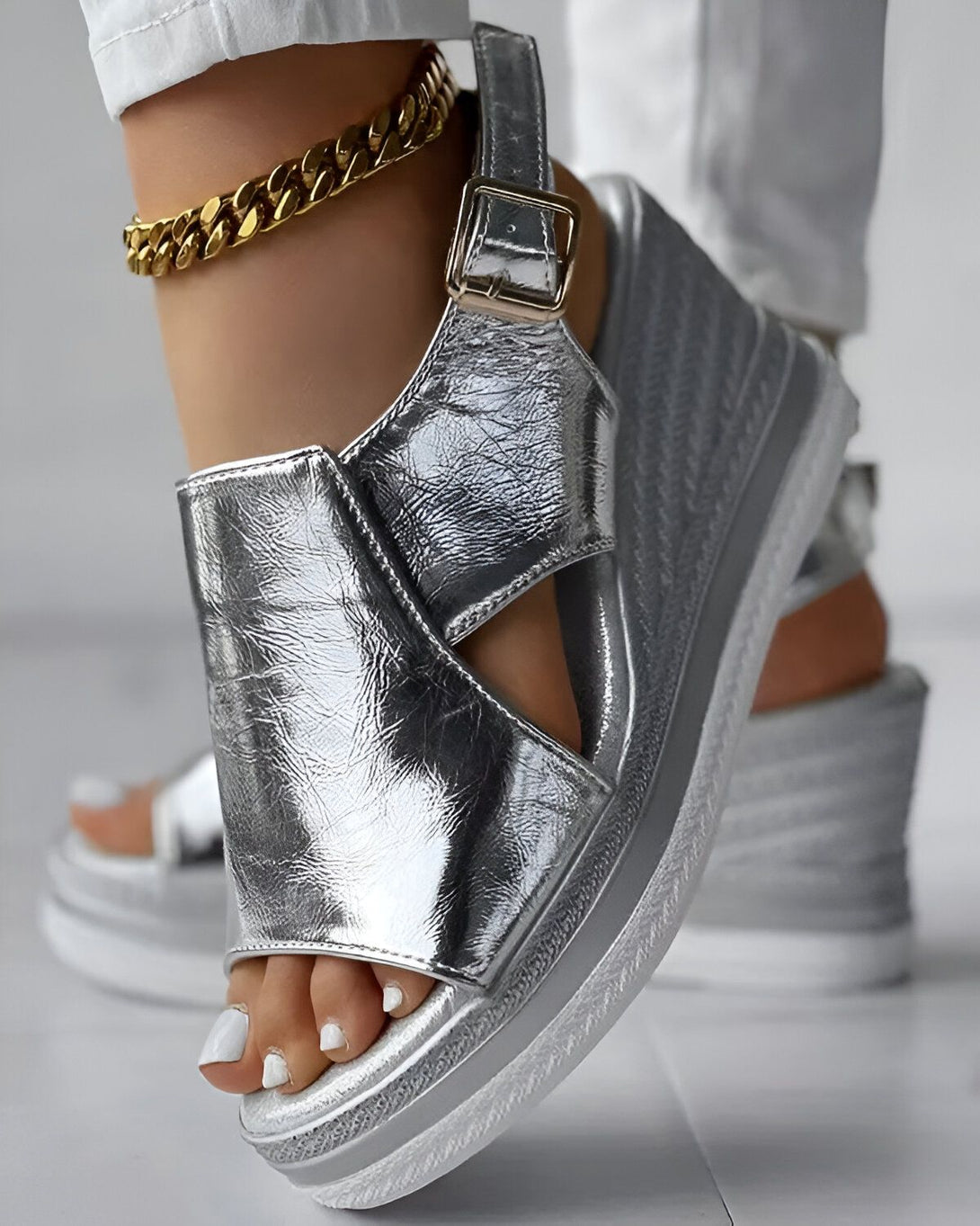 Une femme porte des sandales compensées argentées. Les sandales sont en cuir et ont une bride à boucle. La femme porte un pantalon blanc et un chemisier blanc. Elle a les ongles des pieds peints en blanc.