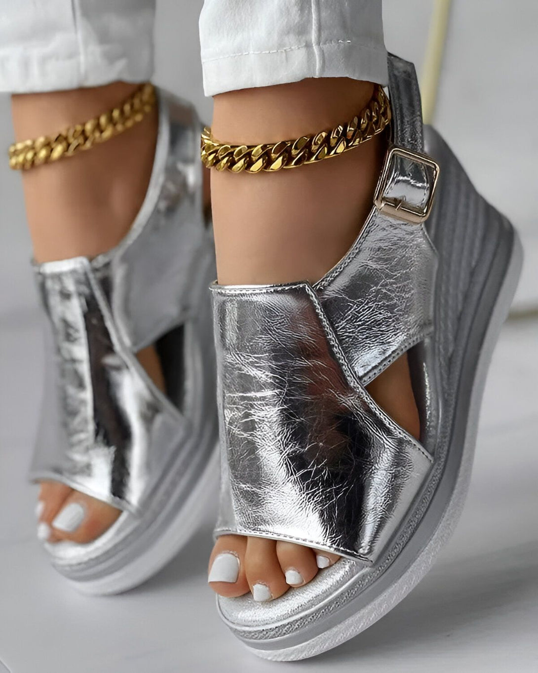 Une femme porte des sandales compensées argentées avec des brides croisées sur le dessus du pied. Elle porte également un pantalon blanc et un bracelet de cheville doré.