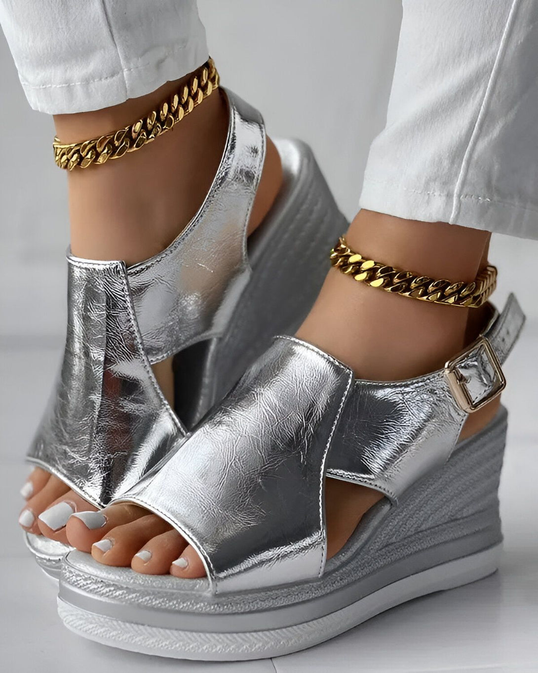 Une femme porte des sandales compensées argentées. Les sandales sont en cuir et ont une bride à boucle. La femme porte également un jean blanc et des bijoux dorés.