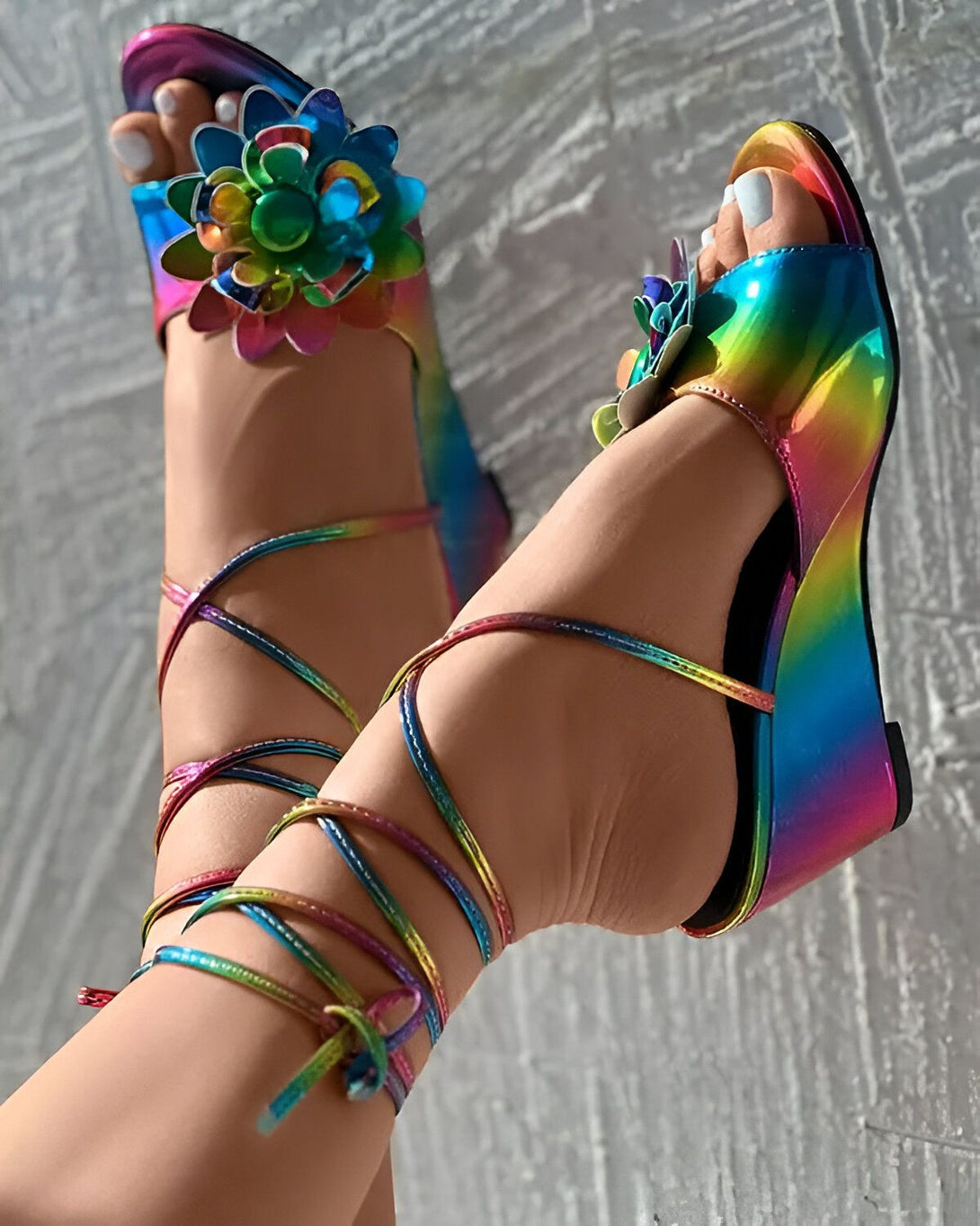 Une paire de sandales compensées arc-en-ciel à brides croisées sur le devant et une fleur en tissu irisé sur le dessus du pied.