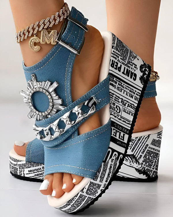 Une femme porte des sandales compensées en jean bleu avec des brides à chaîne et un ornement en strass. Les sandales ont une semelle blanche à motif journal.