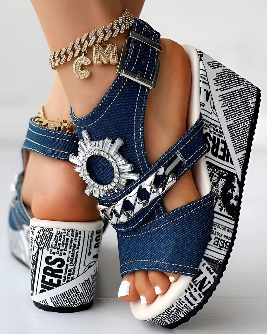 Une femme porte des sandales à talons compensés en jean bleu avec des brides à boucles et des détails cloutés. Les sandales ont une semelle épaisse blanche et marron.