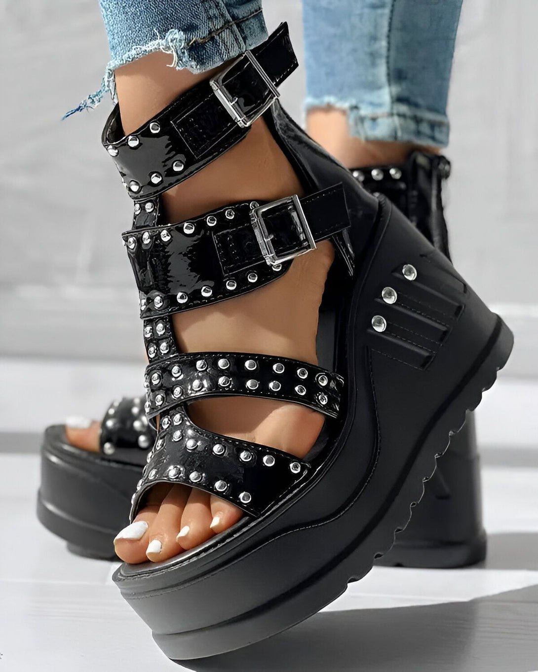 Une femme porte des sandales compensées noires à talons hauts. Les sandales sont ornées de clous argentés. La femme porte un jean bleu et les ongles des pieds peints en blanc.