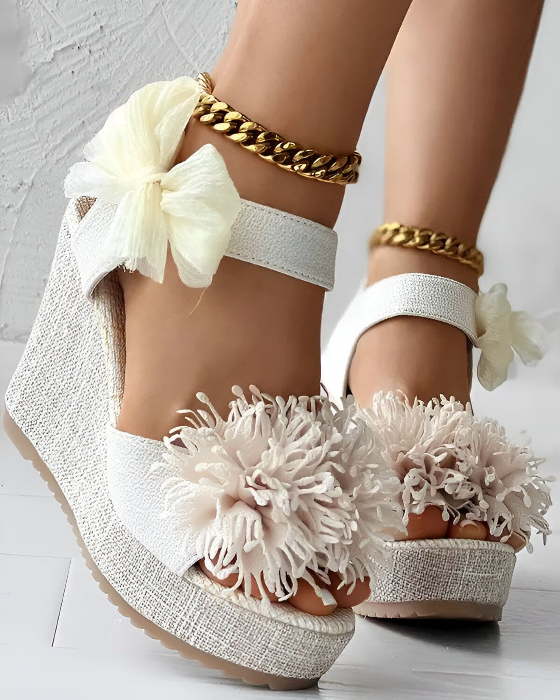 Une paire de sandales compensées blanches à fleurs. Les sandales sont en toile et ont une semelle compensée en corde. Les fleurs sont en tissu et sont de couleur rose pâle. Les sandales sont également dotées d'une bride à la cheville avec une chaîne dorée.