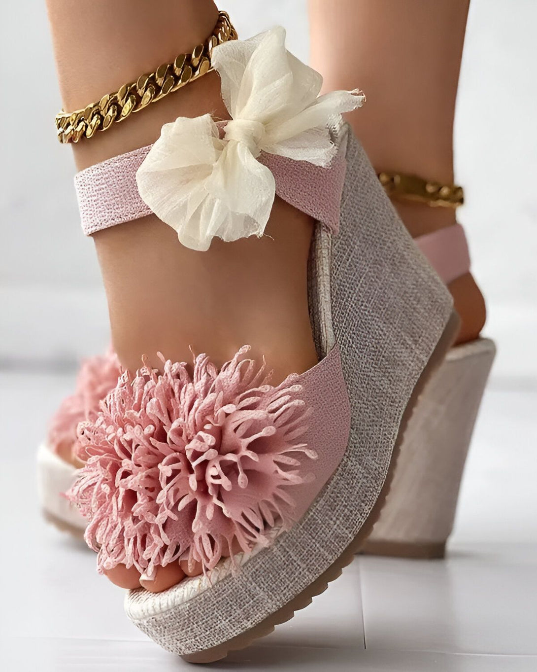 Une femme porte des sandales compensées roses à brides croisées sur le cou-de-pied. Les sandales sont ornées de fleurs en tissu rose pâle. La femme porte également une chaîne dorée autour de la cheville.