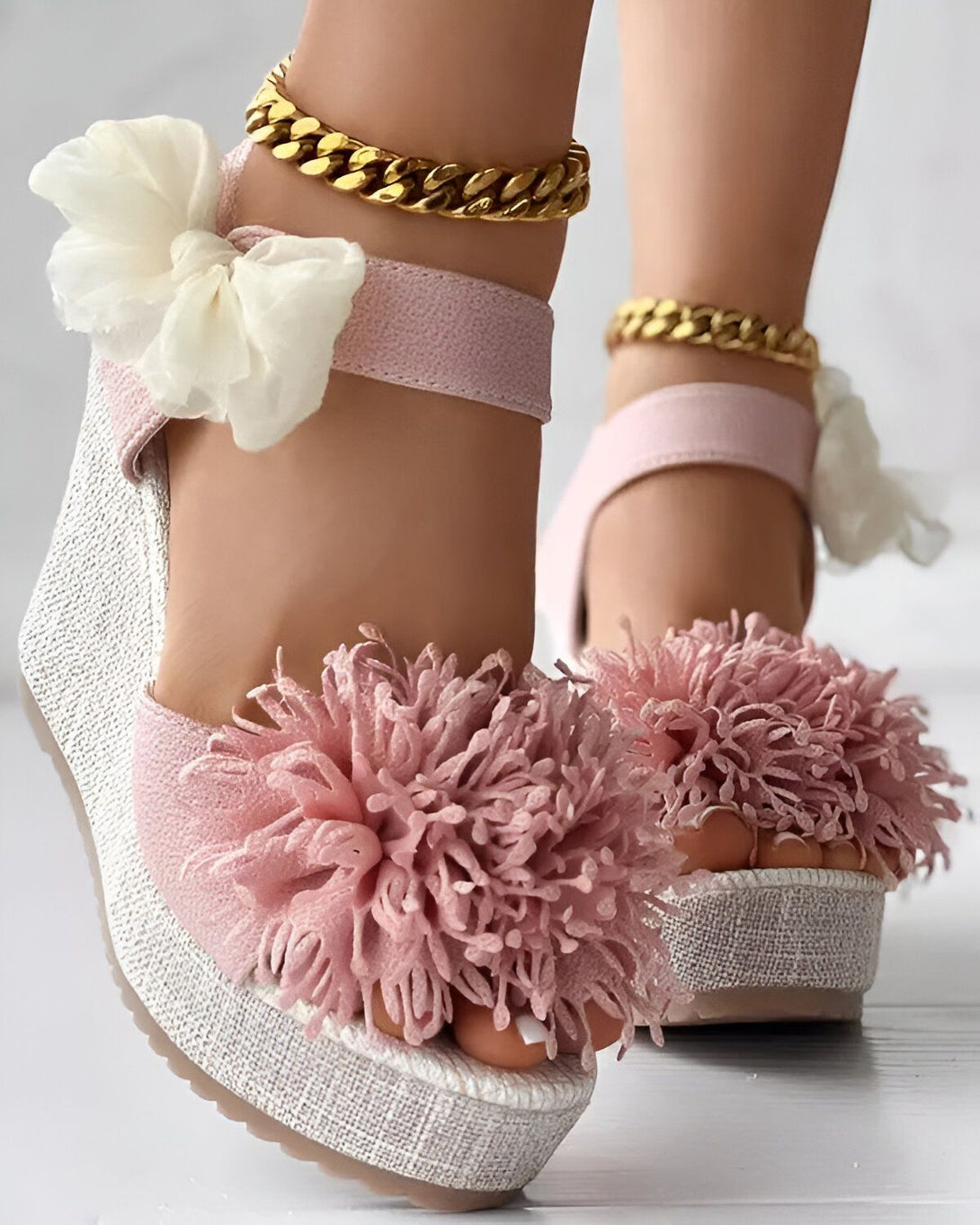 Une paire de sandales compensées roses à brides. Les sandales sont décorées de fleurs en tissu blanc et rose. La semelle est en corde beige et la bride cheville est en chaîne dorée.