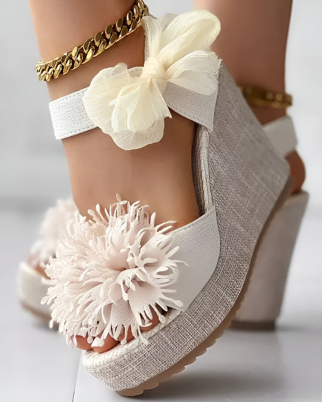 Une femme porte des sandales compensées blanches à brides croisées sur le cou-de-pied. Les sandales sont ornées de fleurs en tissu blanc et rose. La femme porte également une chaîne dorée à la cheville.