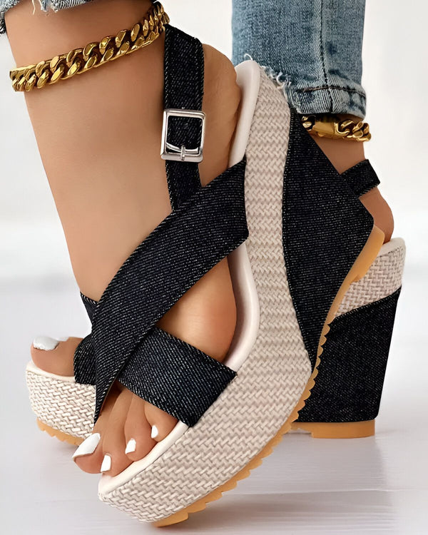Une femme porte des sandales compensées noires et beiges. Les sandales sont en denim et ont une bride croisée sur le dessus du pied. La femme porte également un jean et une cheville dorée.