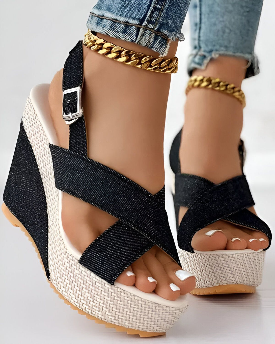 Une femme porte des sandales compensées en denim noir avec des brides croisées sur le dessus du pied. Les sandales ont une semelle compensée en corde beige et une boucle en métal doré sur le côté. La femme porte un jean bleu et un bracelet de cheville en chaîne dorée.