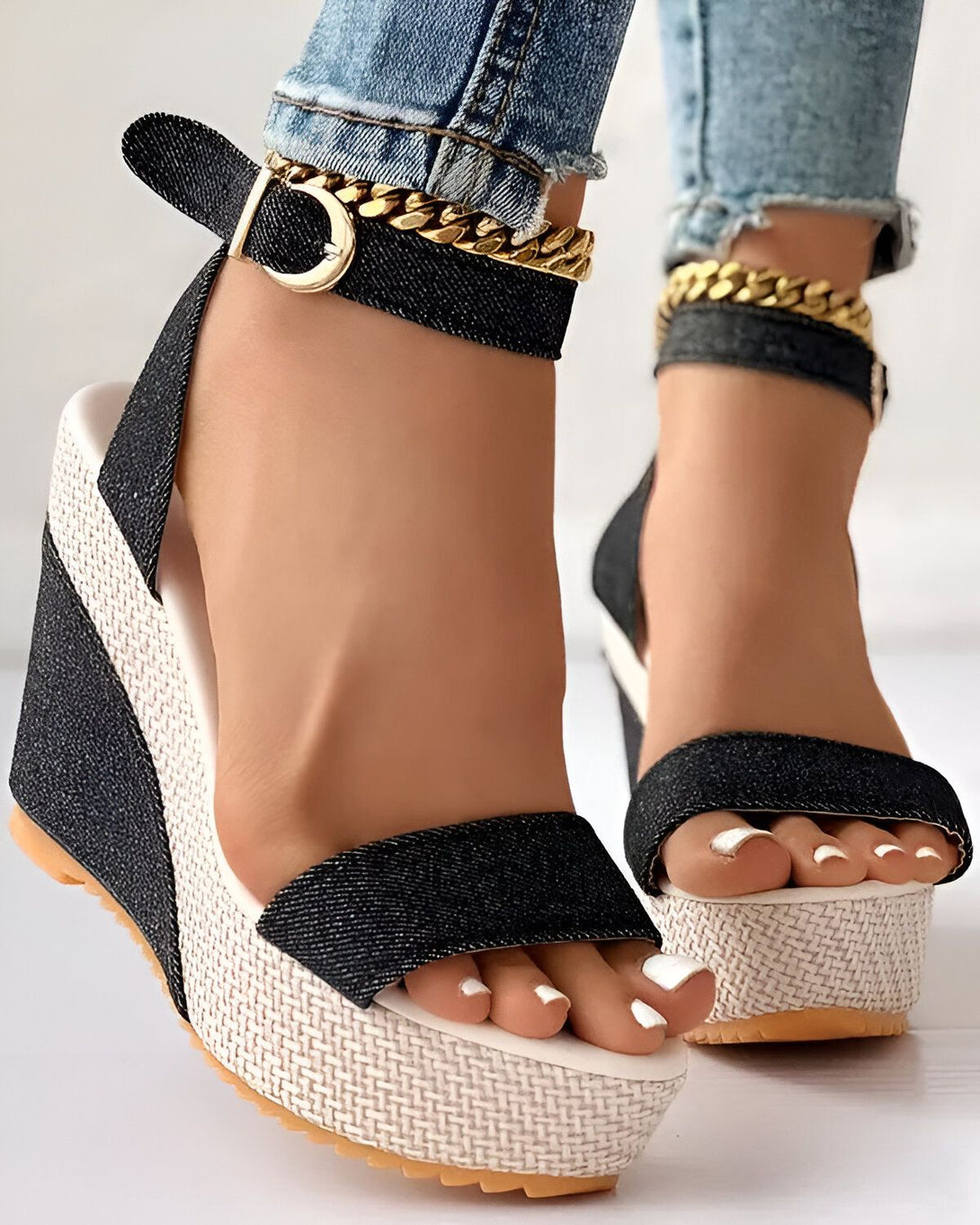 Une femme porte des sandales compensées noires et blanches. Les sandales ont une bride cheville avec une boucle et une chaîne dorée. La femme porte un jean bleu et les ongles des pieds peints en blanc.