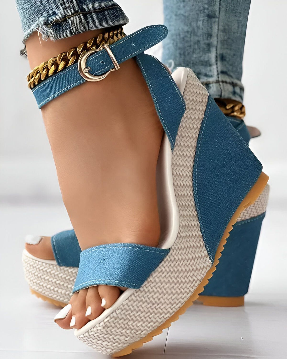 Une femme porte des sandales compensées bleues et blanches. Les sandales ont une bride à la cheville avec une chaîne dorée. La femme porte un jean bleu.