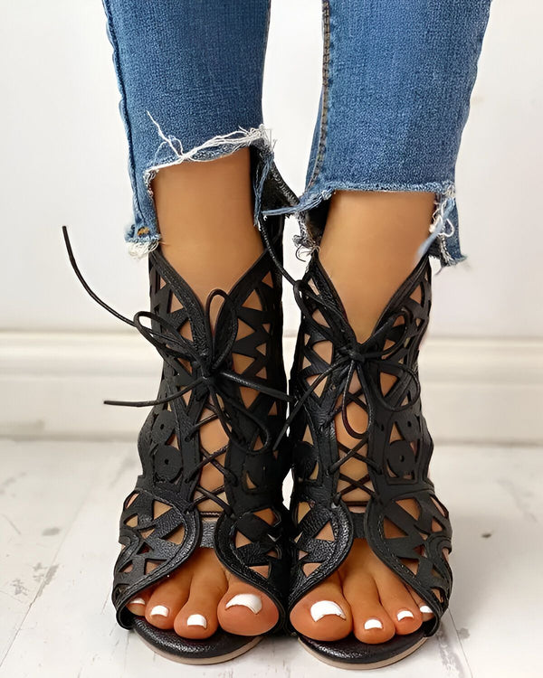 Une femme porte des chaussures noires à talons hauts. Les chaussures sont ouvertes au niveau des orteils et des lacets montent jusqu'à la cheville. La femme porte un jean bleu déchiré au niveau des genoux.