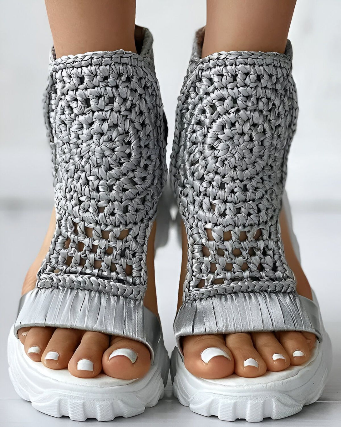 Une paire de sandales argentées avec un talon compensé et des brides en crochet. Les sandales sont portées par une femme qui a les ongles des pieds peints en blanc.