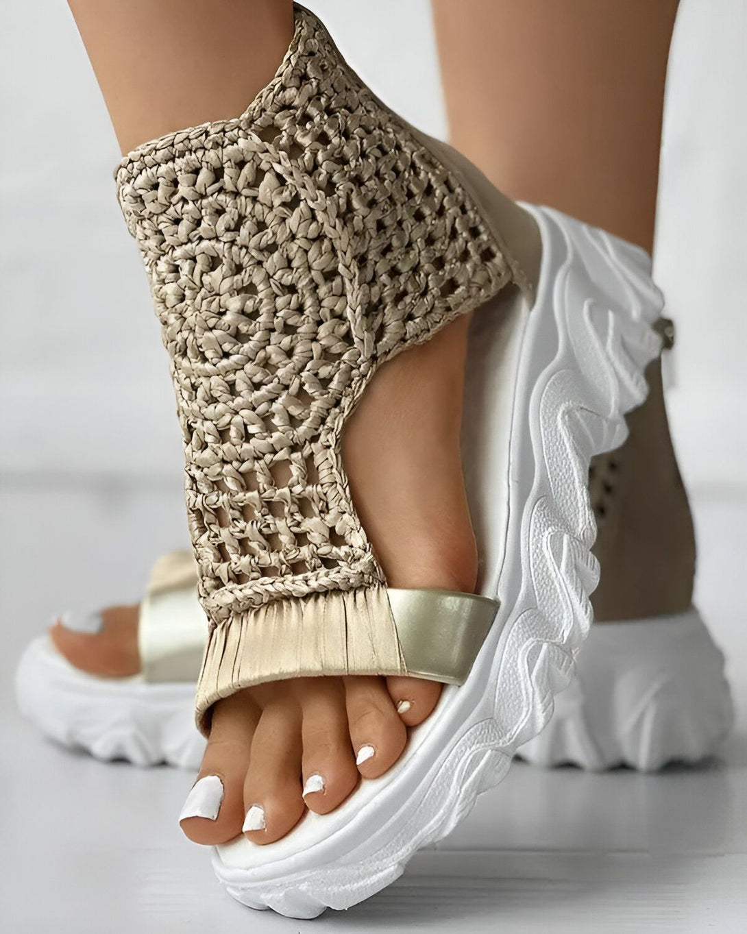 Une femme porte des sandales compensées beiges et blanches. Les sandales sont en crochet et ont une bride à scratch. La semelle est blanche et épaisse.
