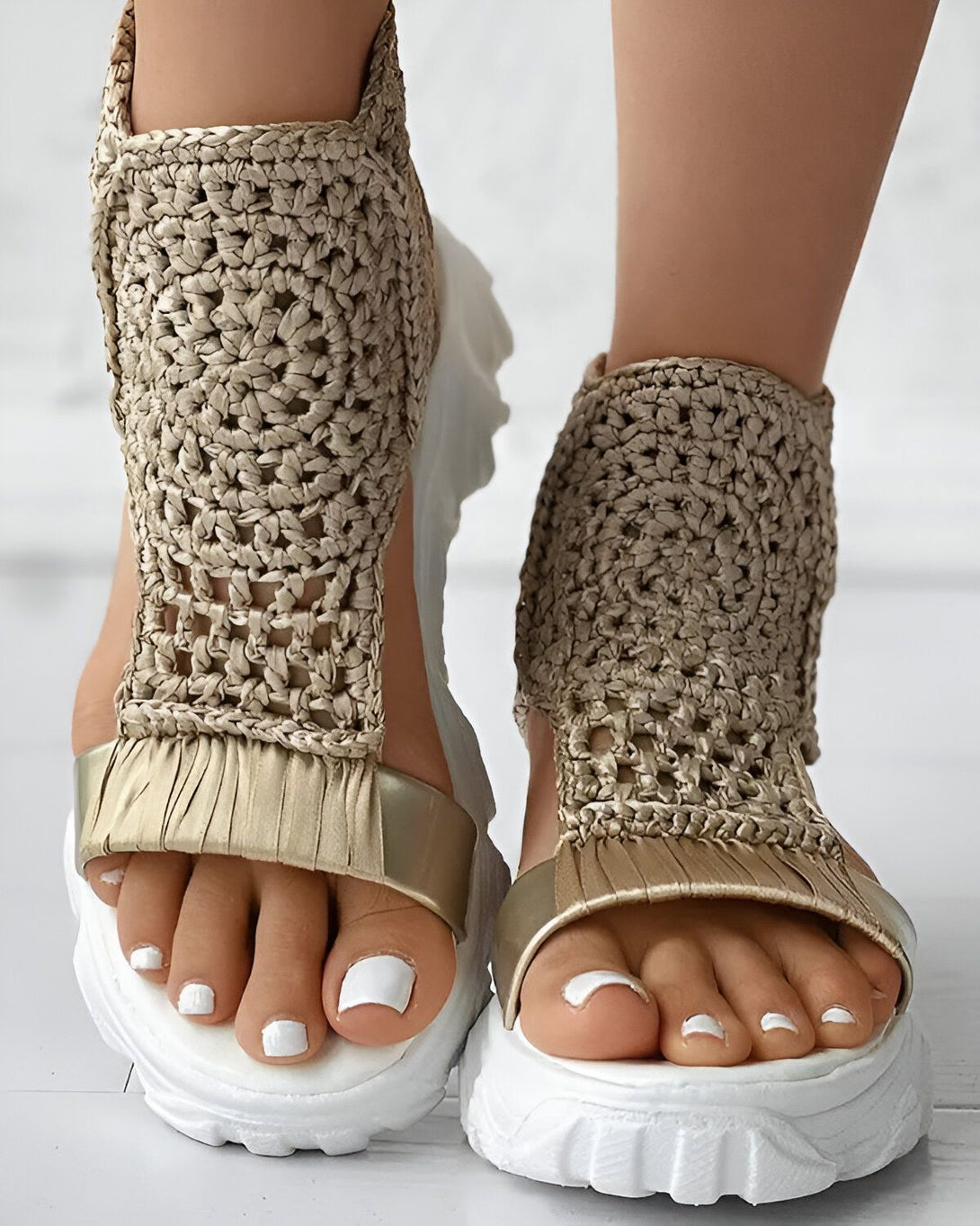 Une femme porte des sandales plates dorées avec des brides en crochet. Les sandales ont une semelle blanche épaisse et les orteils sont blancs.