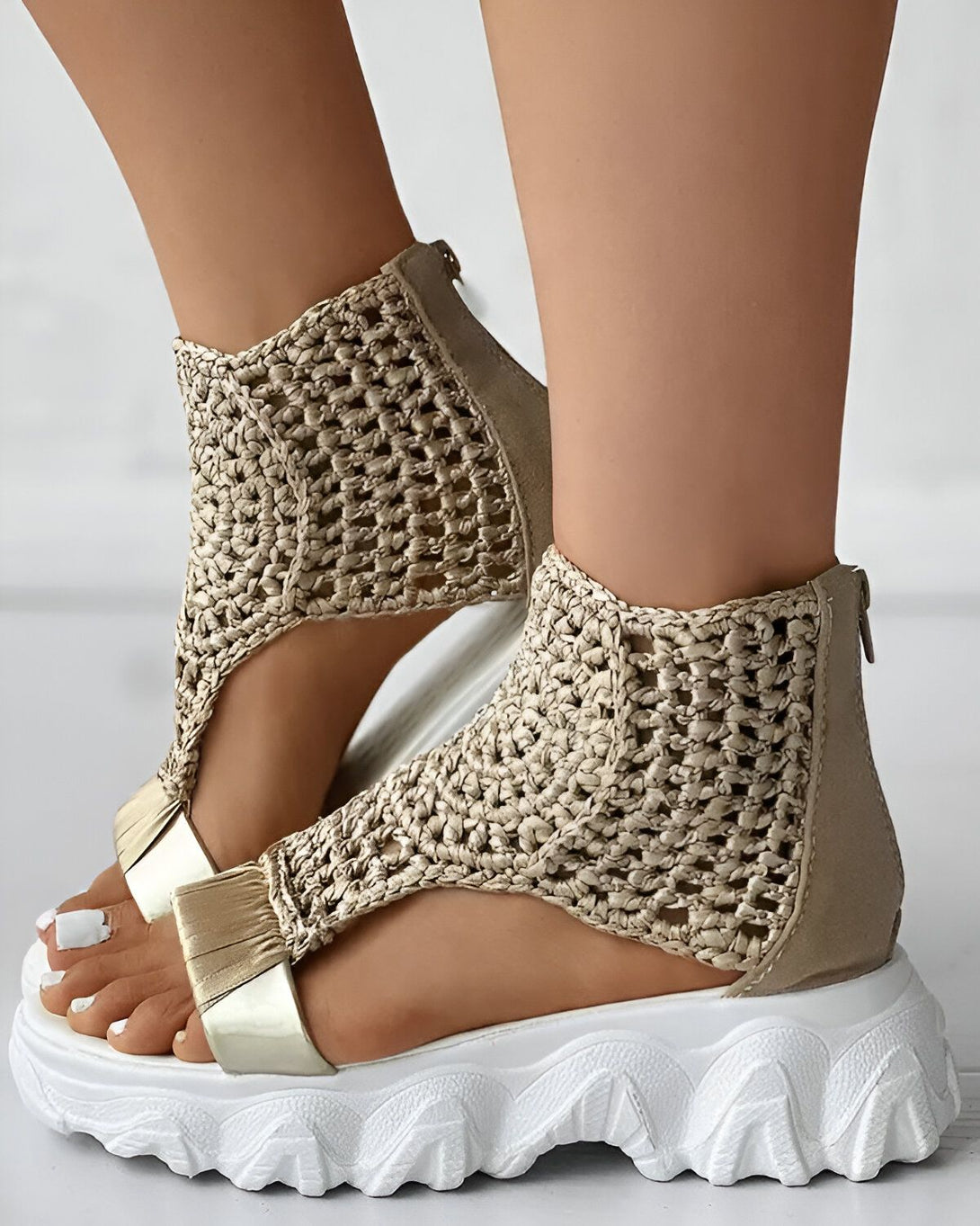 Une femme porte des sandales compensées beiges et blanches. Les sandales sont en matière synthétique et ont un talon de 5 cm. Elles sont dotées d'une fermeture éclair sur le côté pour un enfilage facile.