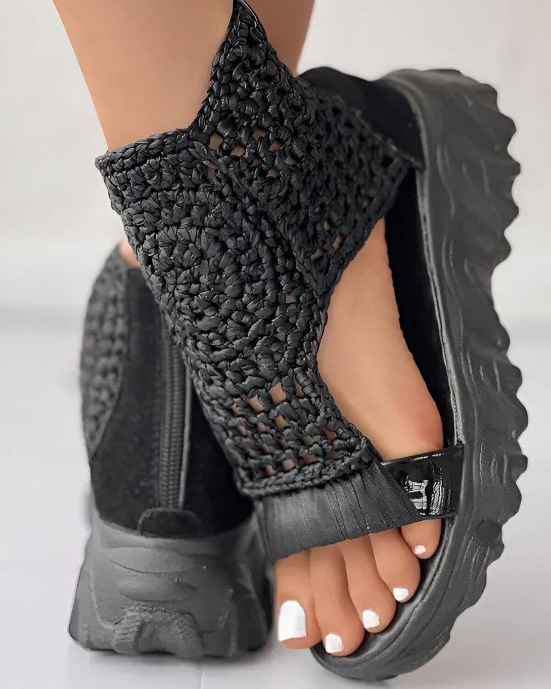 Une femme porte des sandales noires à talons compensés. Les sandales sont en cuir et ont un bout ouvert. La femme a les ongles des pieds peints en blanc.
