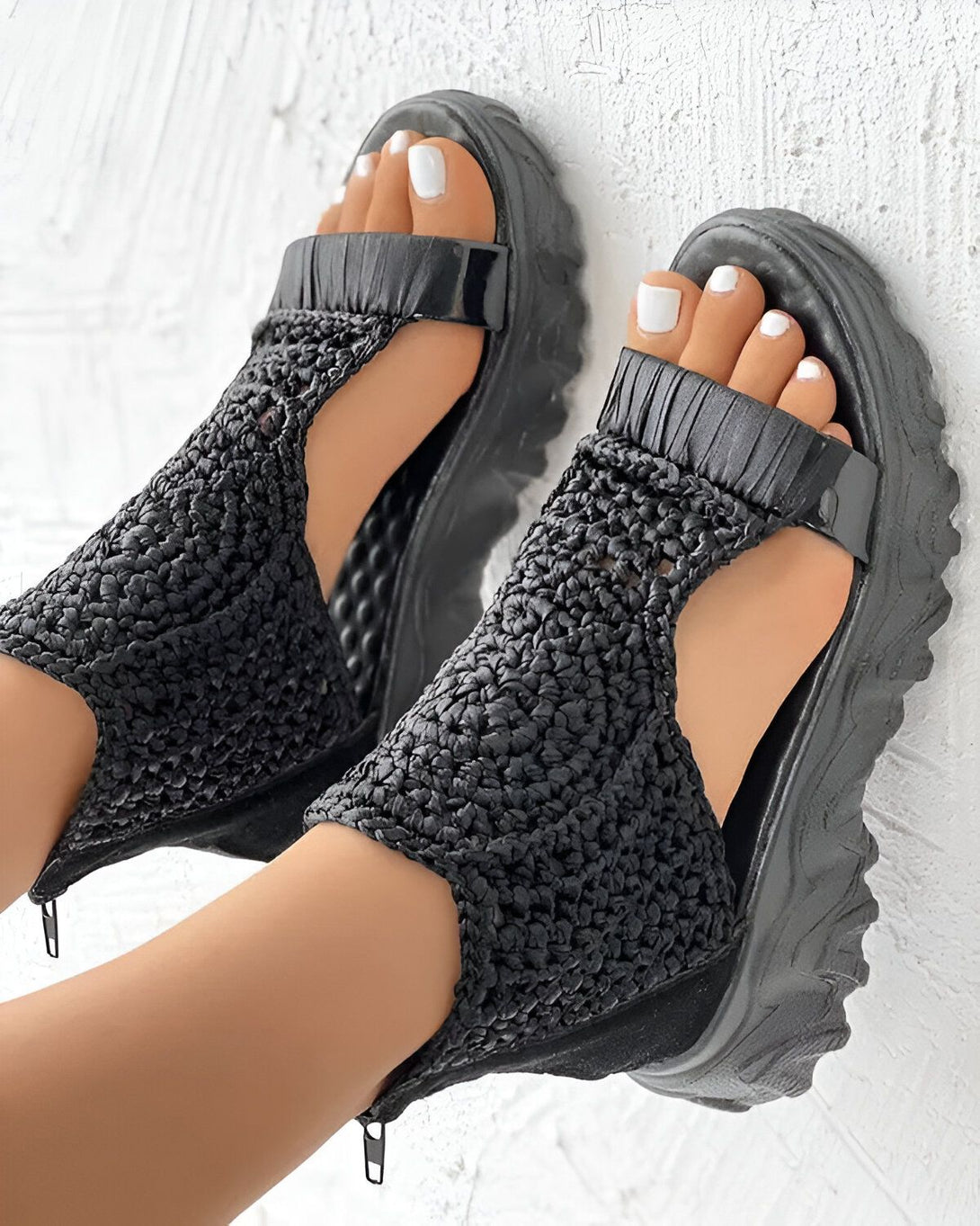 Une paire de sandales noires à talons compensés avec des brides en crochet. Les sandales sont portées par une femme qui a les ongles des pieds peints en blanc.