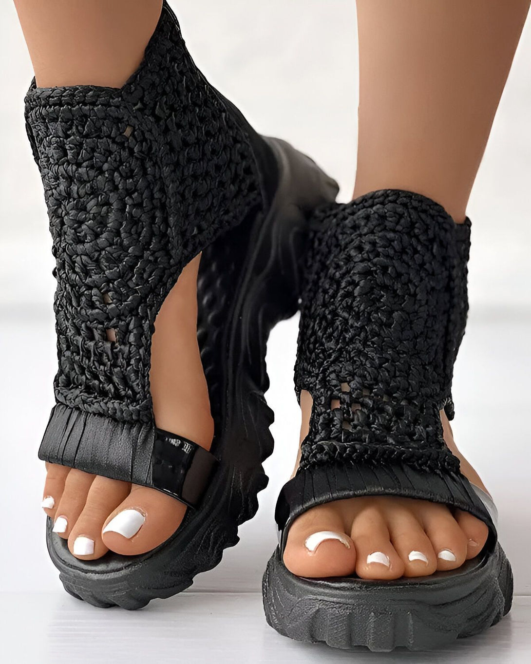 Une femme porte des sandales noires avec des semelles épaisses. Les sandales sont faites de lanières de cuir tressées et ont des brides qui s'attachent autour de la cheville. La femme a les ongles des pieds peints en blanc.