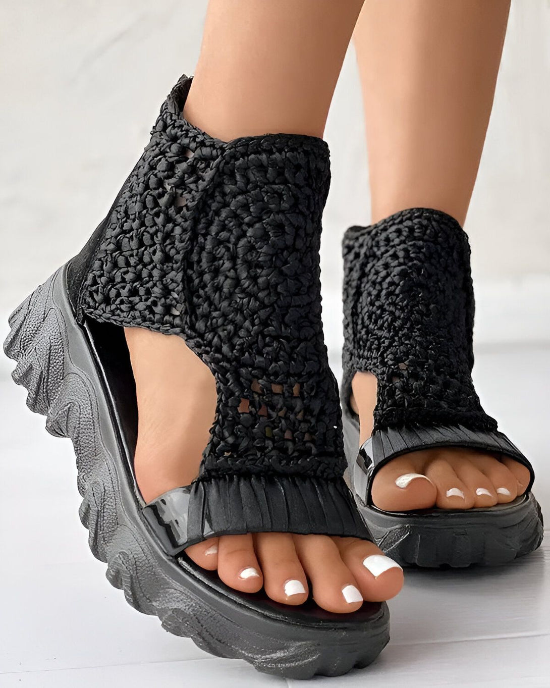 Une femme porte des sandales noires à talons compensés. Les sandales sont en crochet et ont une bride qui s'enroule autour de la cheville. La femme a les ongles des pieds peints en blanc.