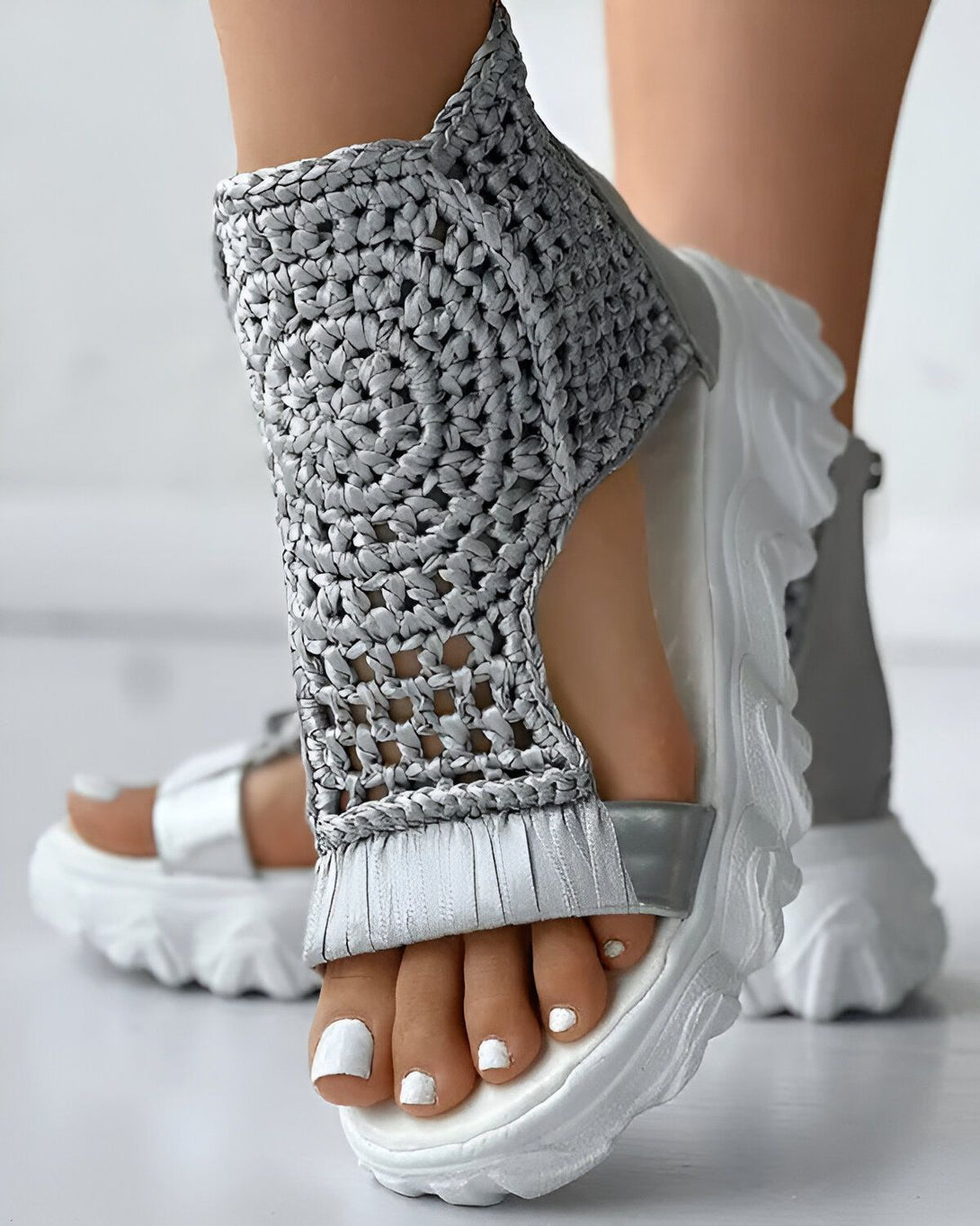 Une femme porte des sandales grises à talons compensés. Les sandales sont en matière tressée et ont des brides qui s'attachent au niveau de la cheville. La femme a les ongles des pieds peints en blanc.