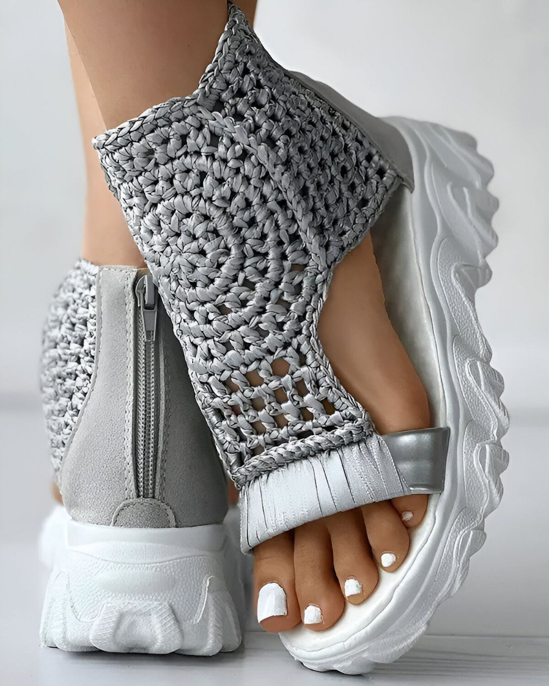 Une femme porte des sandales grises à talons compensés. Les sandales sont en maille et ont une fermeture éclair sur le côté. La femme a les ongles des pieds peints en blanc.