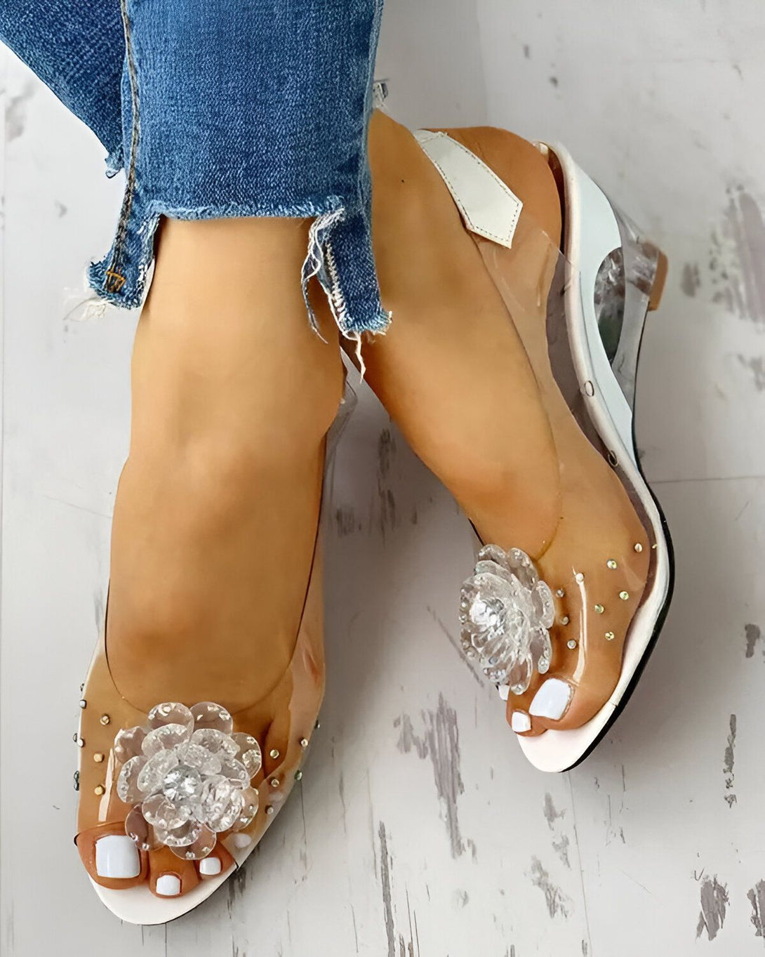 Une femme porte des sandales à talons compensés transparentes avec des fleurs en strass. Elle porte un jean bleu déchiré.